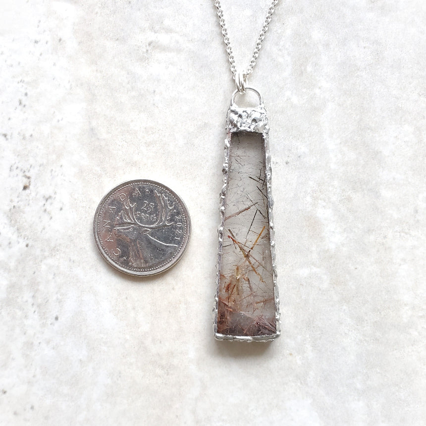 Silver pendant with a textured design next to a coin on a light gray background