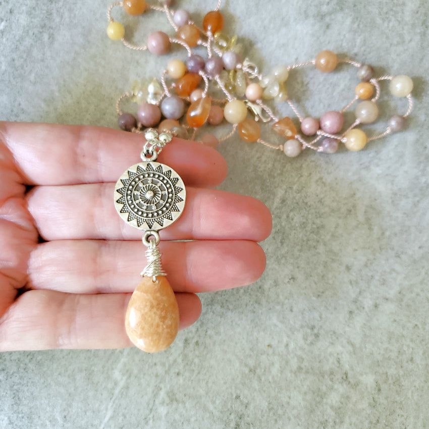 Necklace with a sun-shaped pendant and multi coloured gemstones held by a hand on a light background