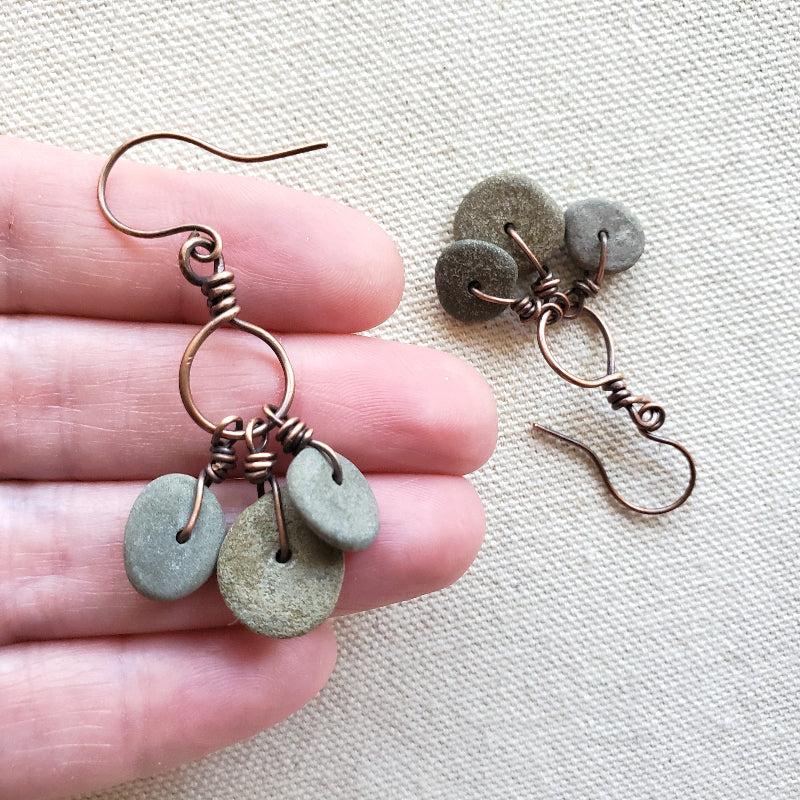 Hand holding a pair of earrings with green stones and copper wire on a beige fabric background