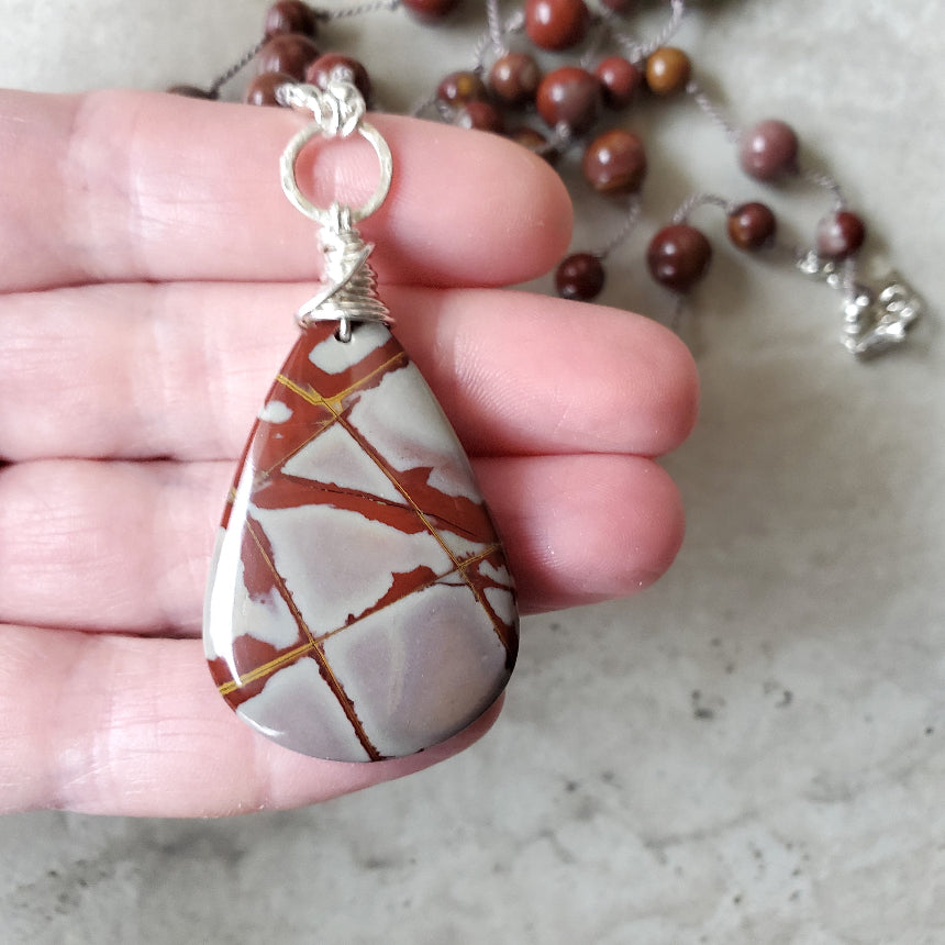 Hand holding a teardrop-shaped stone pendant with a silver wire wrap, with a blurred background of beads.