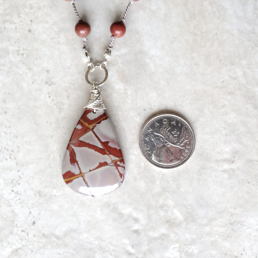 Teardrop-shaped pendant with marbled design on a white textured background, next to a coin for scale.