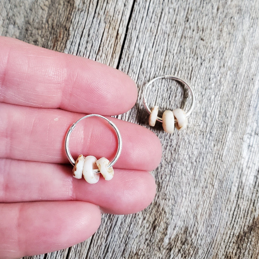 Silver hoop earrings with natural shell beads held between fingers on a wooden surface