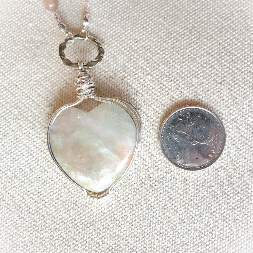 Silver pendant with a heart-shaped stone next to a Canadian dollar coin on a textured fabric background