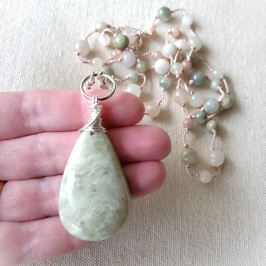 Hand holding a light green teardrop pendant with a beaded necklace on a beige background