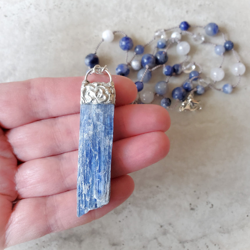 Blue Kyanite pendant in hand with knotted necklace  on tile
