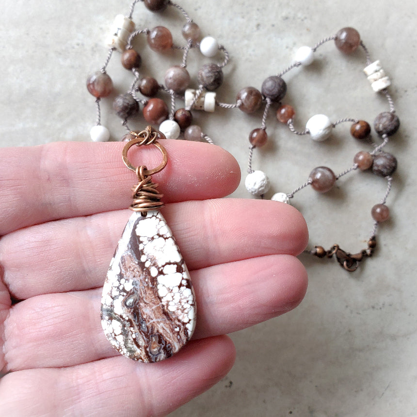 Hand holding a teardrop-shaped pendant with a textured stone design, necklace in the background.